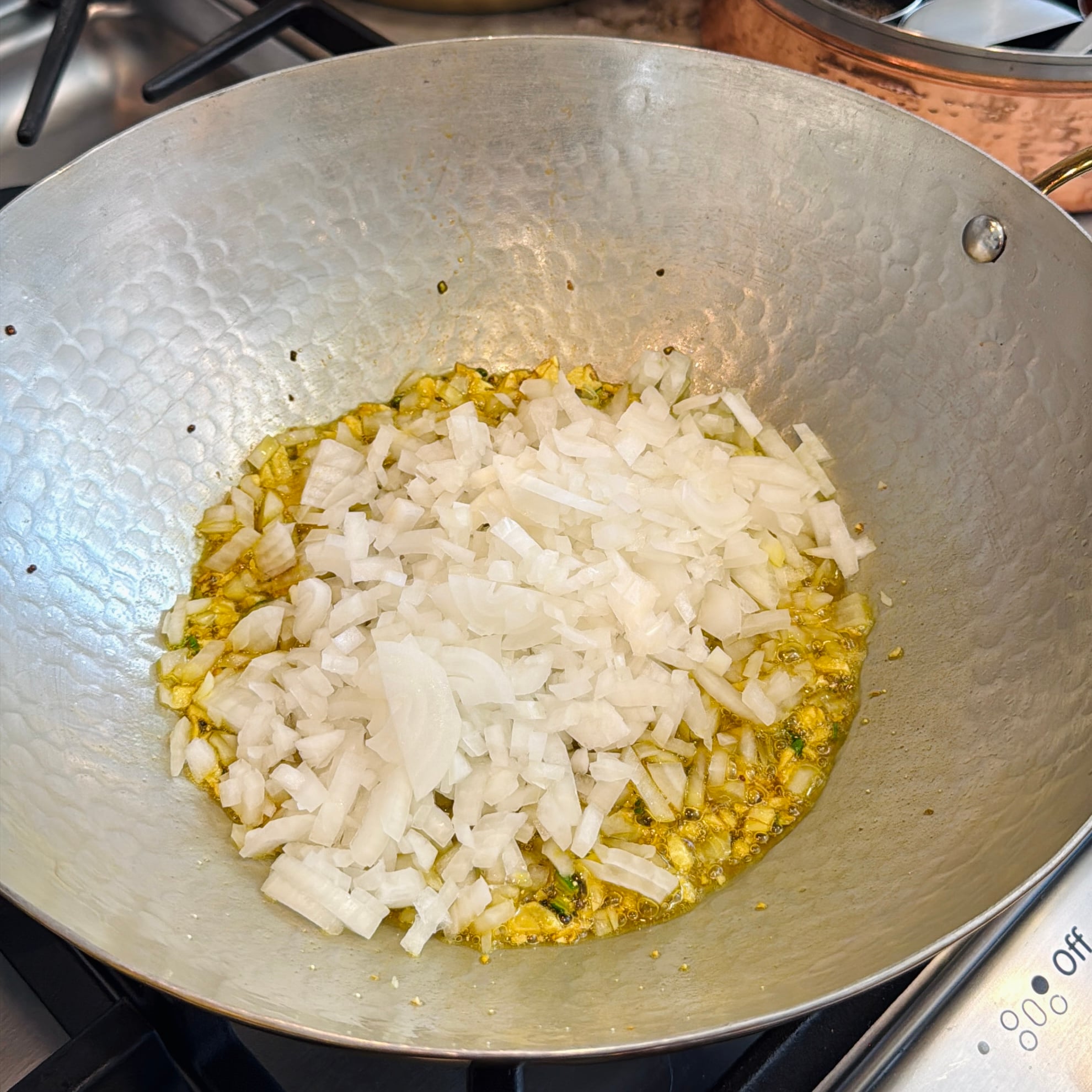Chopped white onions cooking in hot oil with spices in a metal wok on a stovetop.