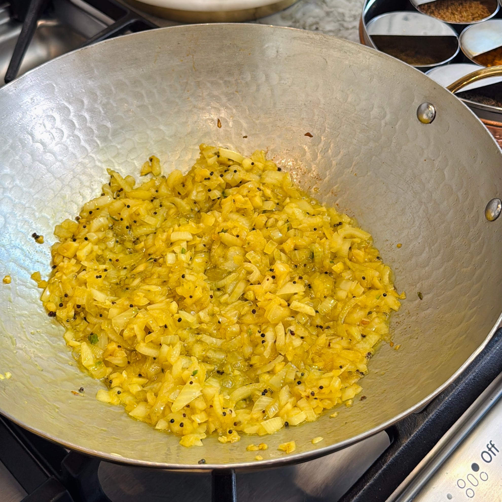 Chopped onions sautéing with spices in a large metal pan on a stovetop; spice box visible in the background.