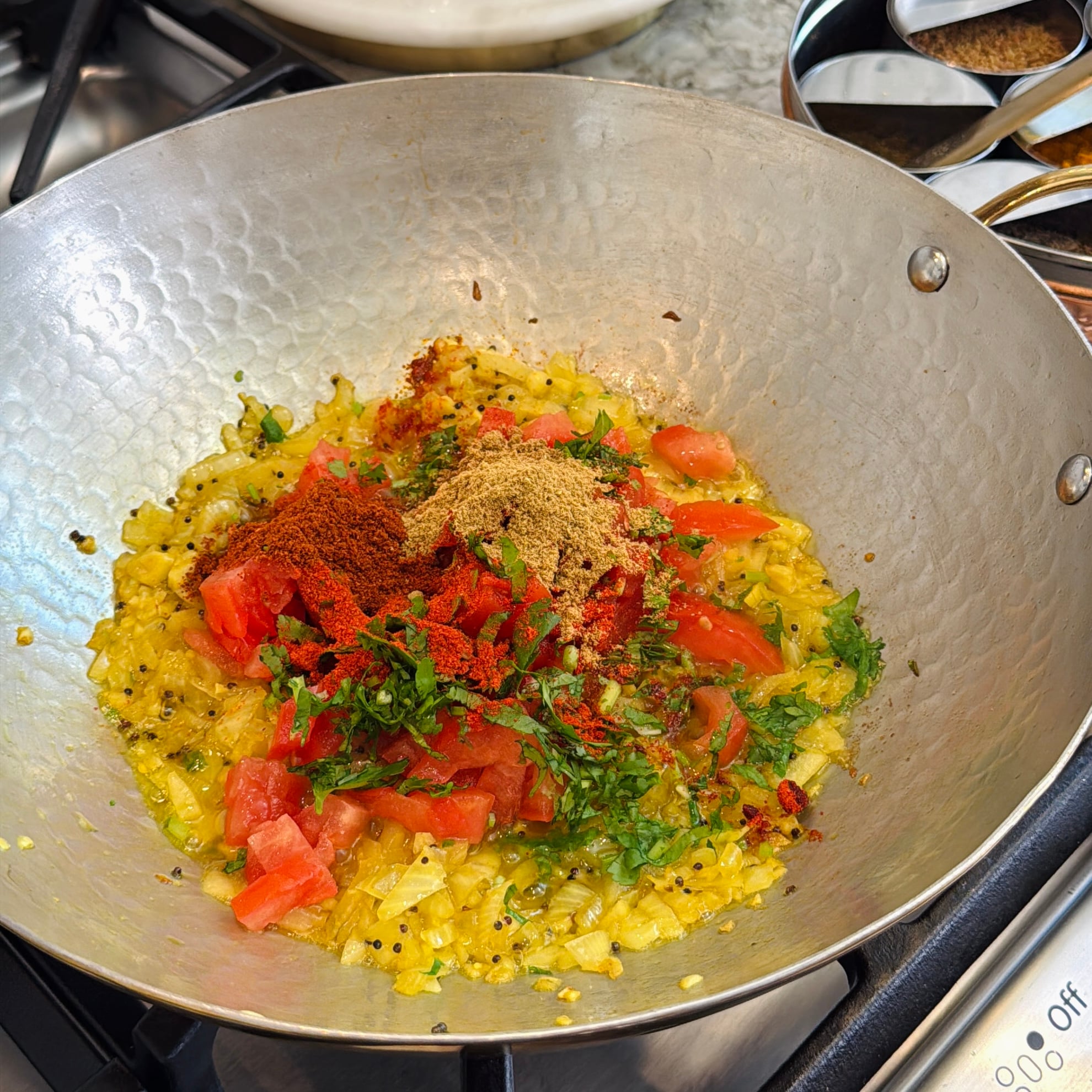Chopped onions, tomatoes, herbs, and spices are cooking in a metal pan on a stovetop. Multiple spice containers are visible in the background.