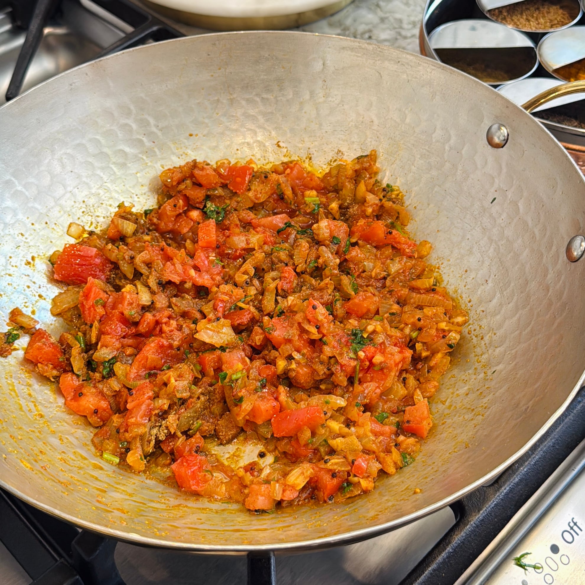 Chopped onions and tomatoes with spices cooking in a metal pan on a stovetop, with a spice container visible in the background.