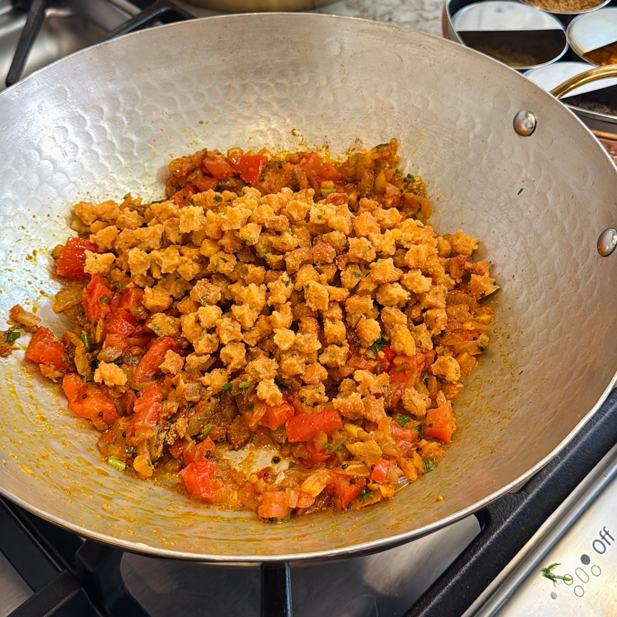 A metal pan on a stove contains chopped tomatoes, onions, and fried sandge.