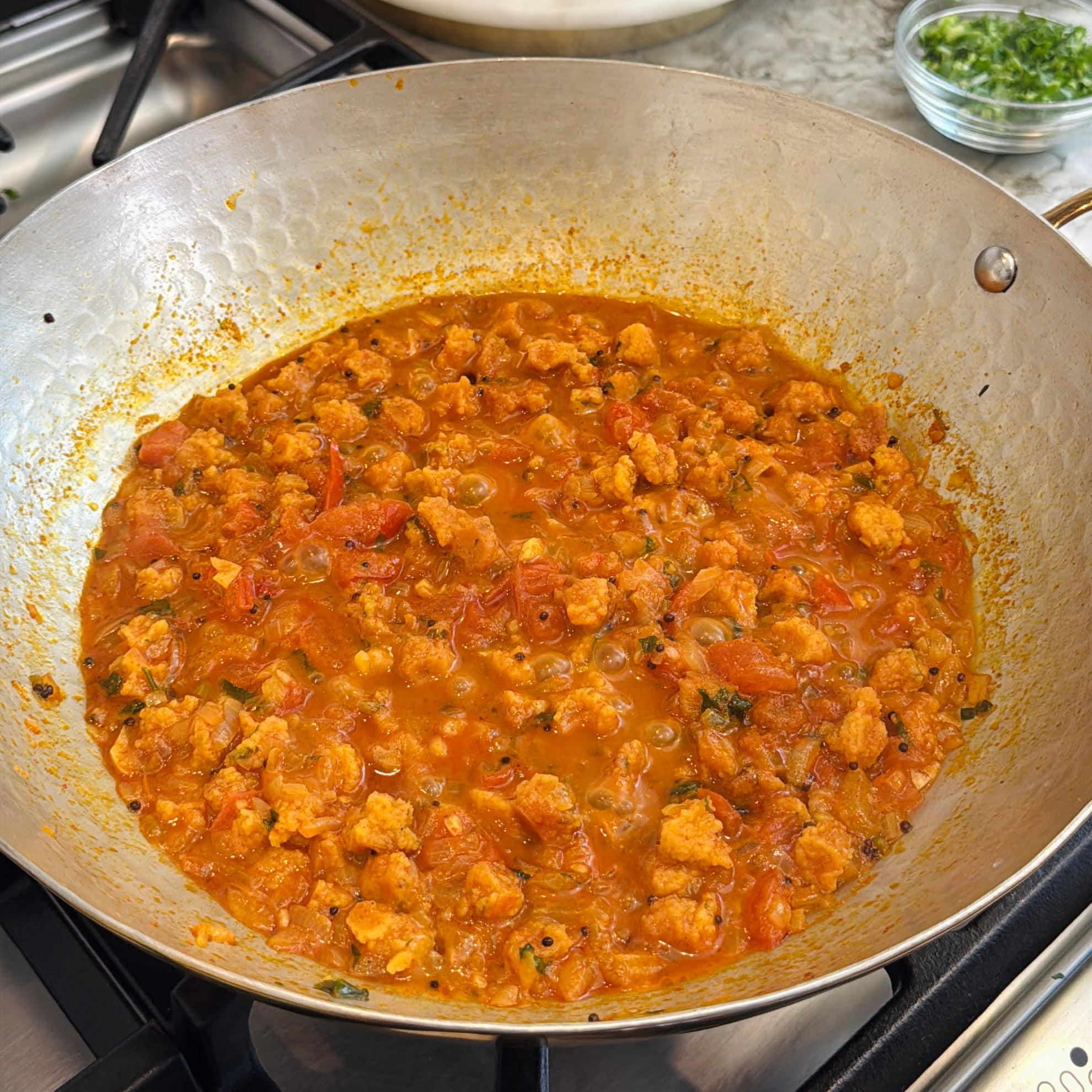 A metal pan on a stovetop contains a thick, orange-red curry with sandge. A small bowl of chopped herbs sits nearby.