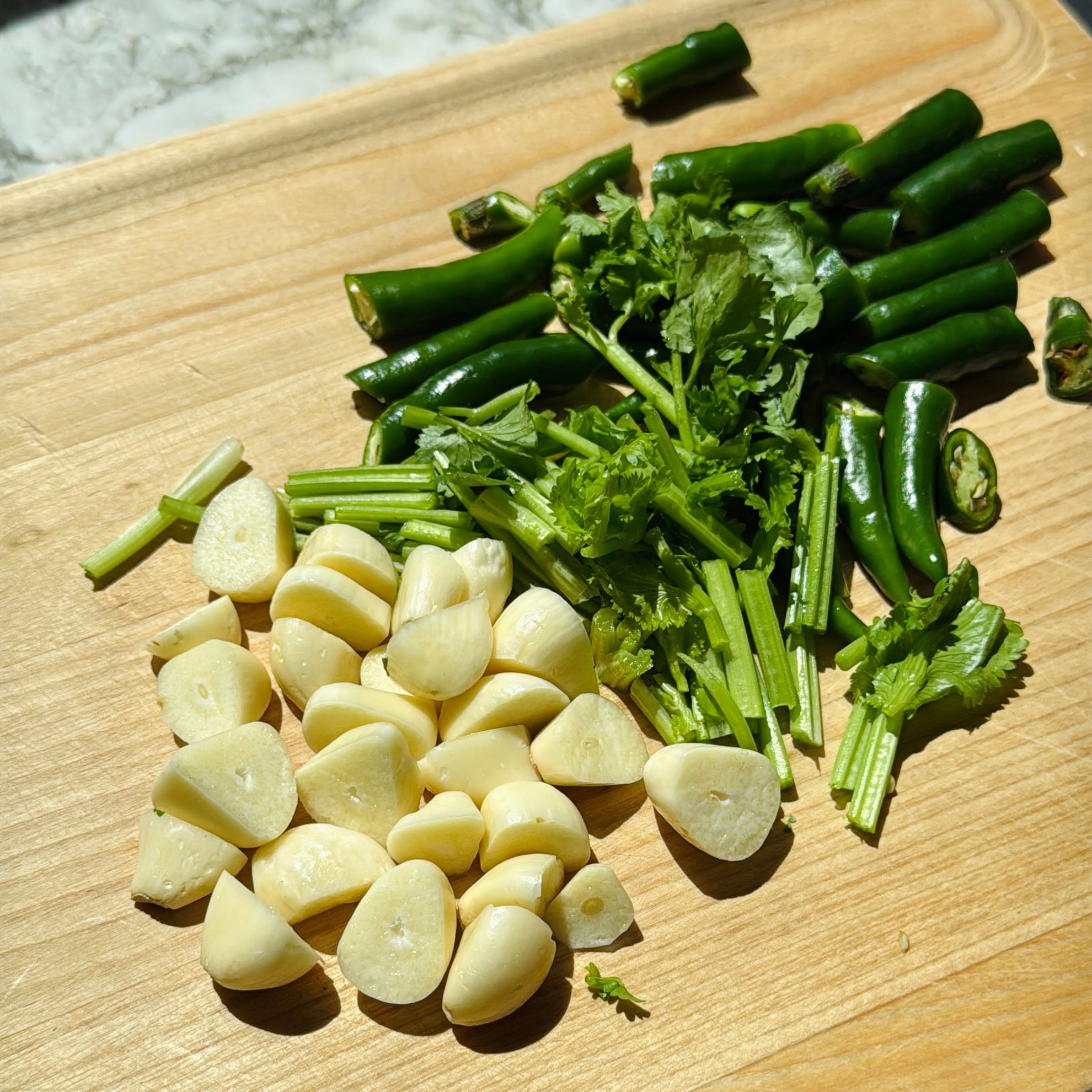 Chopped garlic cloves, fresh cilantro, and sliced green chilies on a wooden cutting board in sunlight.