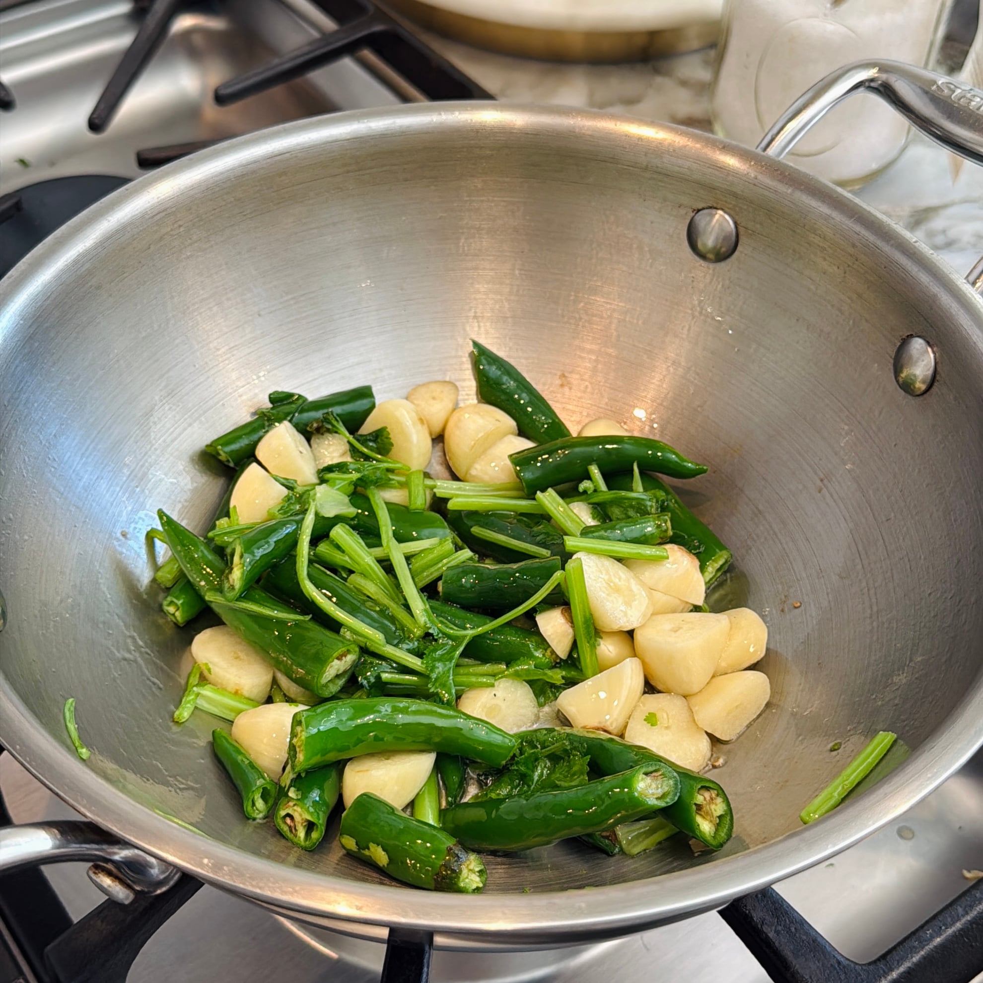 Whole garlic cloves, green chili peppers, and herb stems in a stainless steel pan on a stove, ready to cook.
