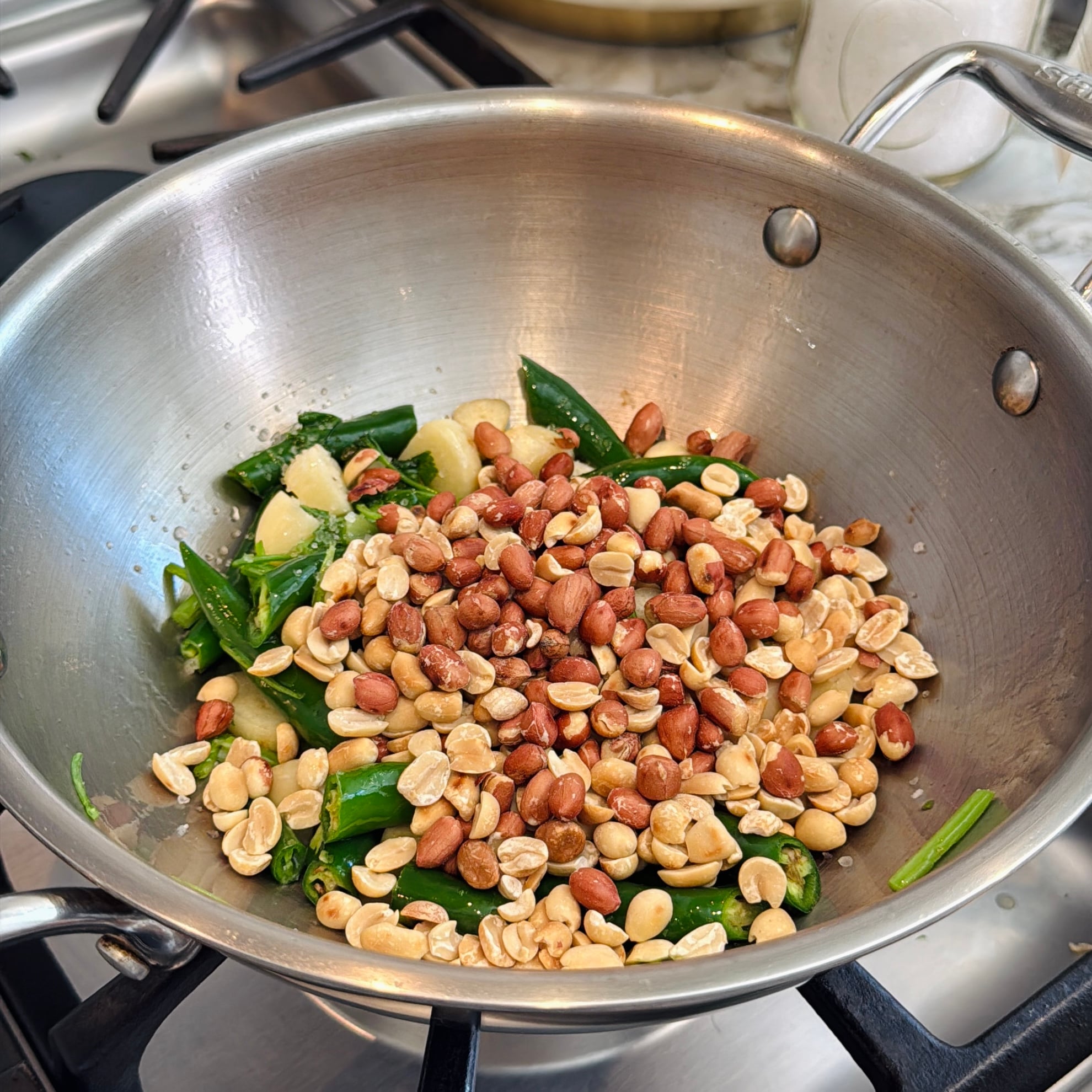 A stainless steel pan on a stove contains chopped green chilies, garlic cloves, and raw peanuts being sautéed together.