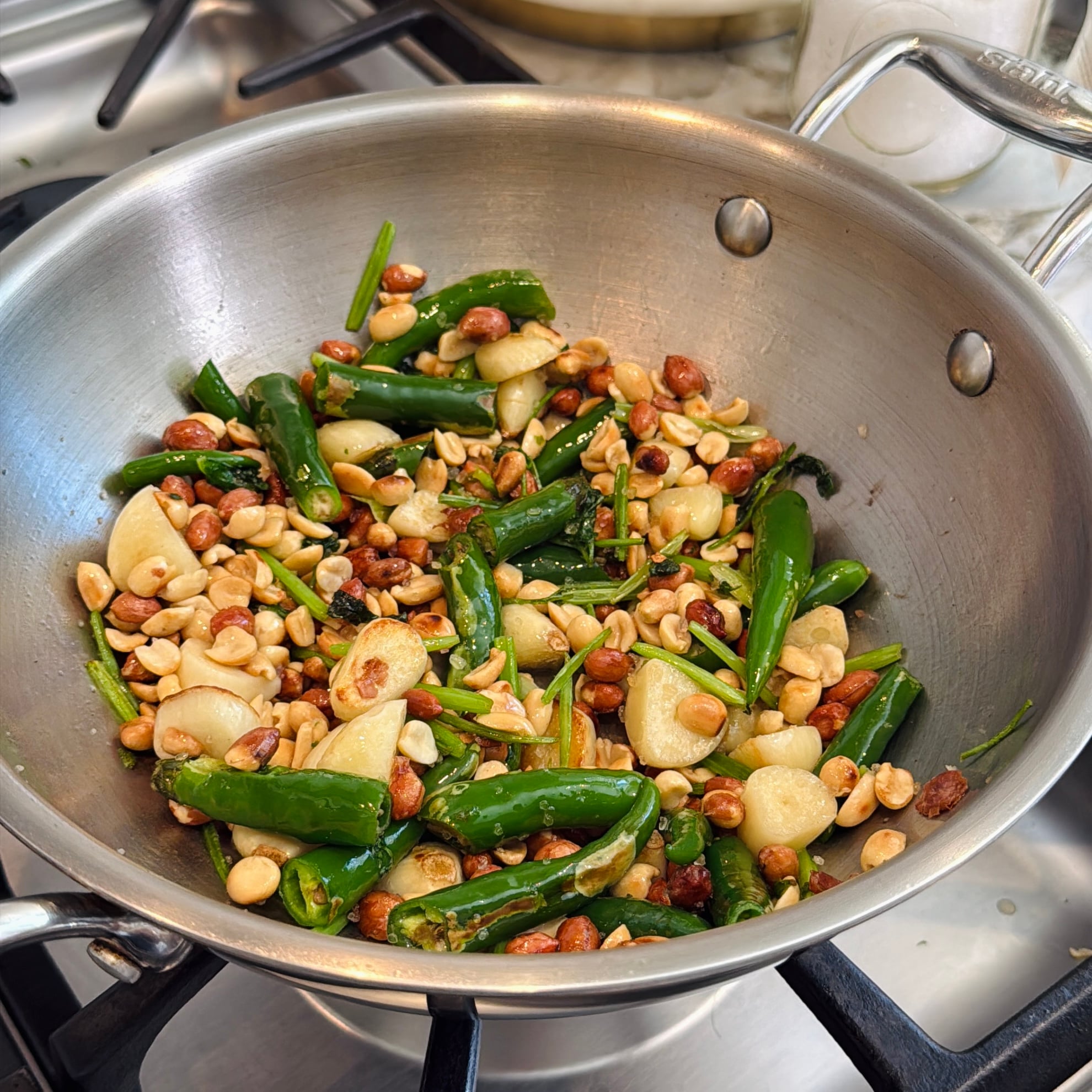 Stainless steel pan on stove containing stir-fried green chilies, garlic cloves, peanuts, and some chopped herbs.