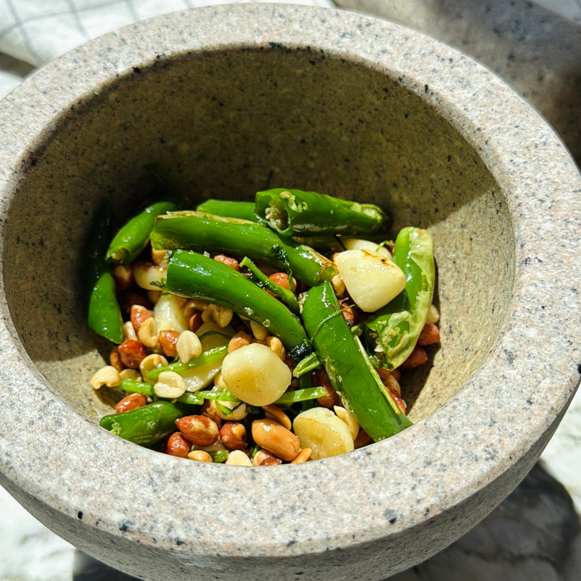 A stone bowl containing green chilies, garlic cloves, roasted peanuts, and chopped herbs, under bright natural light.