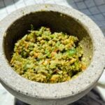 A stone mortar filled with a coarse, green herb and nut mixture, possibly a freshly made pesto or chutney, sitting on a light surface.
