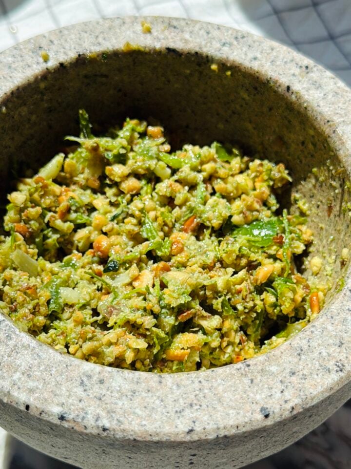 A stone mortar filled with a coarse, green herb and nut mixture, possibly a freshly made pesto or chutney, sitting on a light surface.