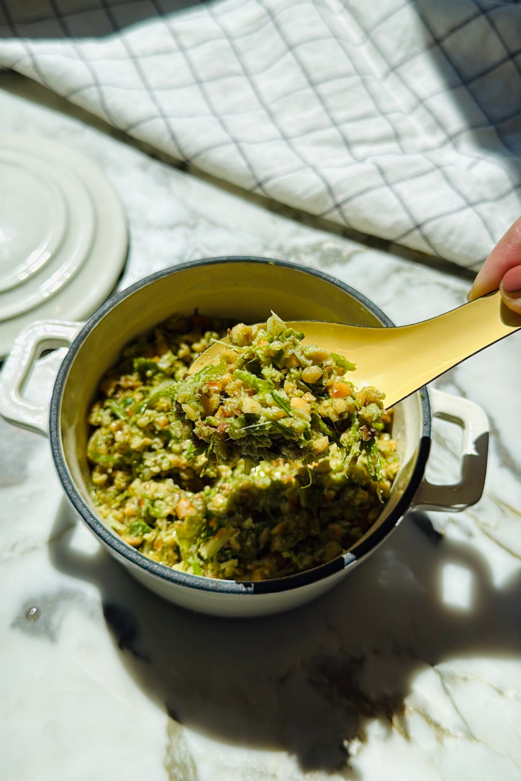 A yellow spoon scoops a chunky green and beige mixture from a white pot on a marble countertop with a folded cloth nearby.
