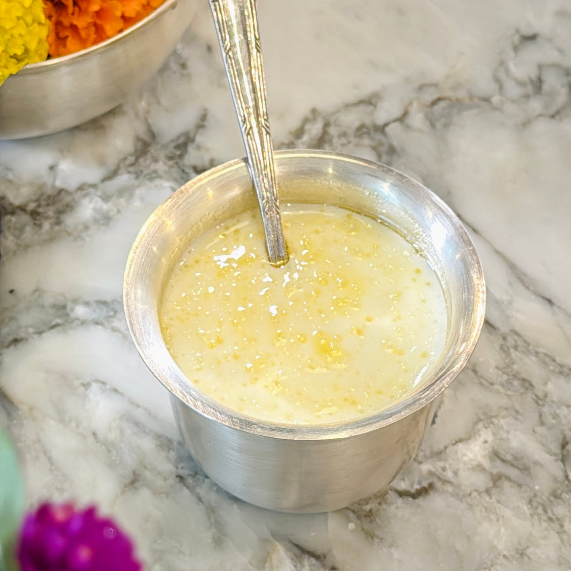 A metal cup filled with a light-colored, grainy liquid, possibly a traditional Indian sweet dish, sits on a marble surface with a spoon inside.