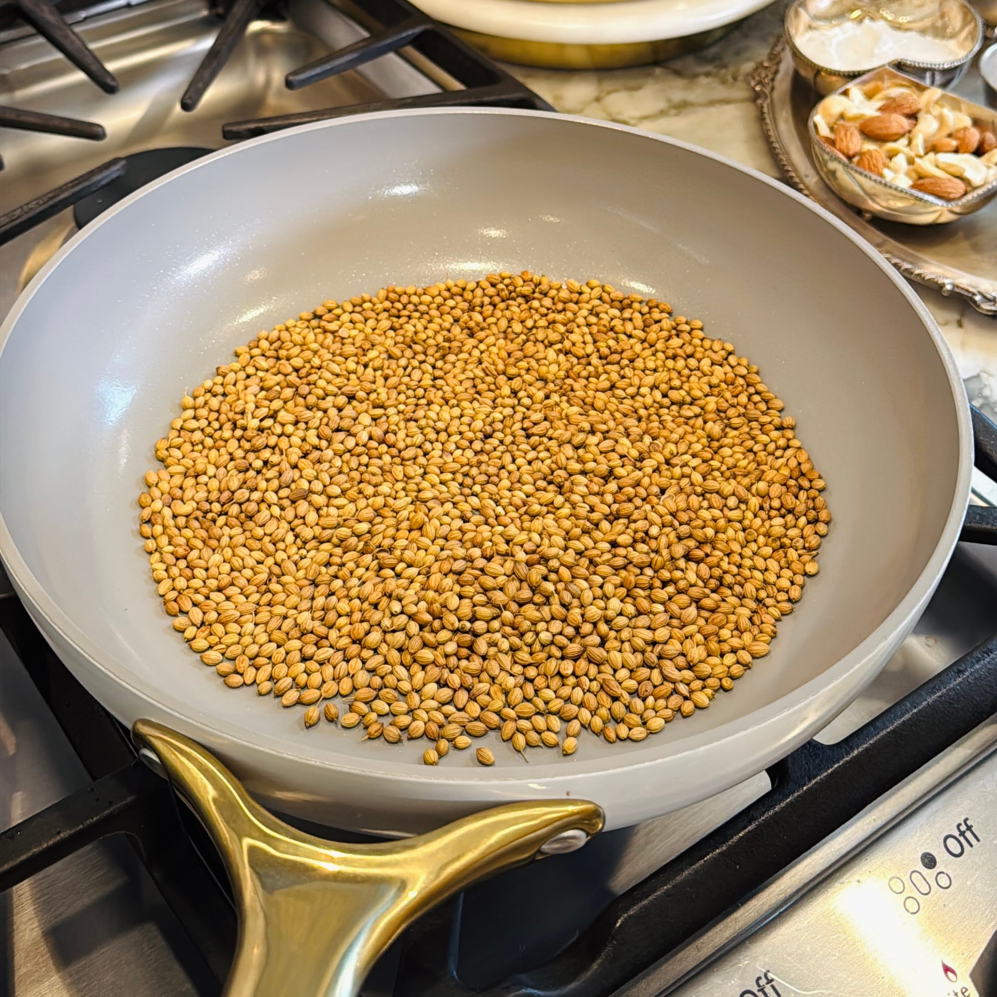 A gray skillet with coriander seeds toasting on a stovetop burner.