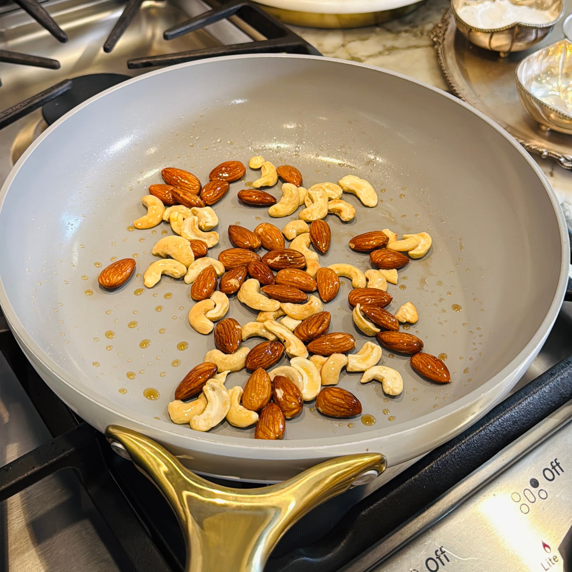 Almonds and cashews are toasting in a gray nonstick frying pan on a stovetop.