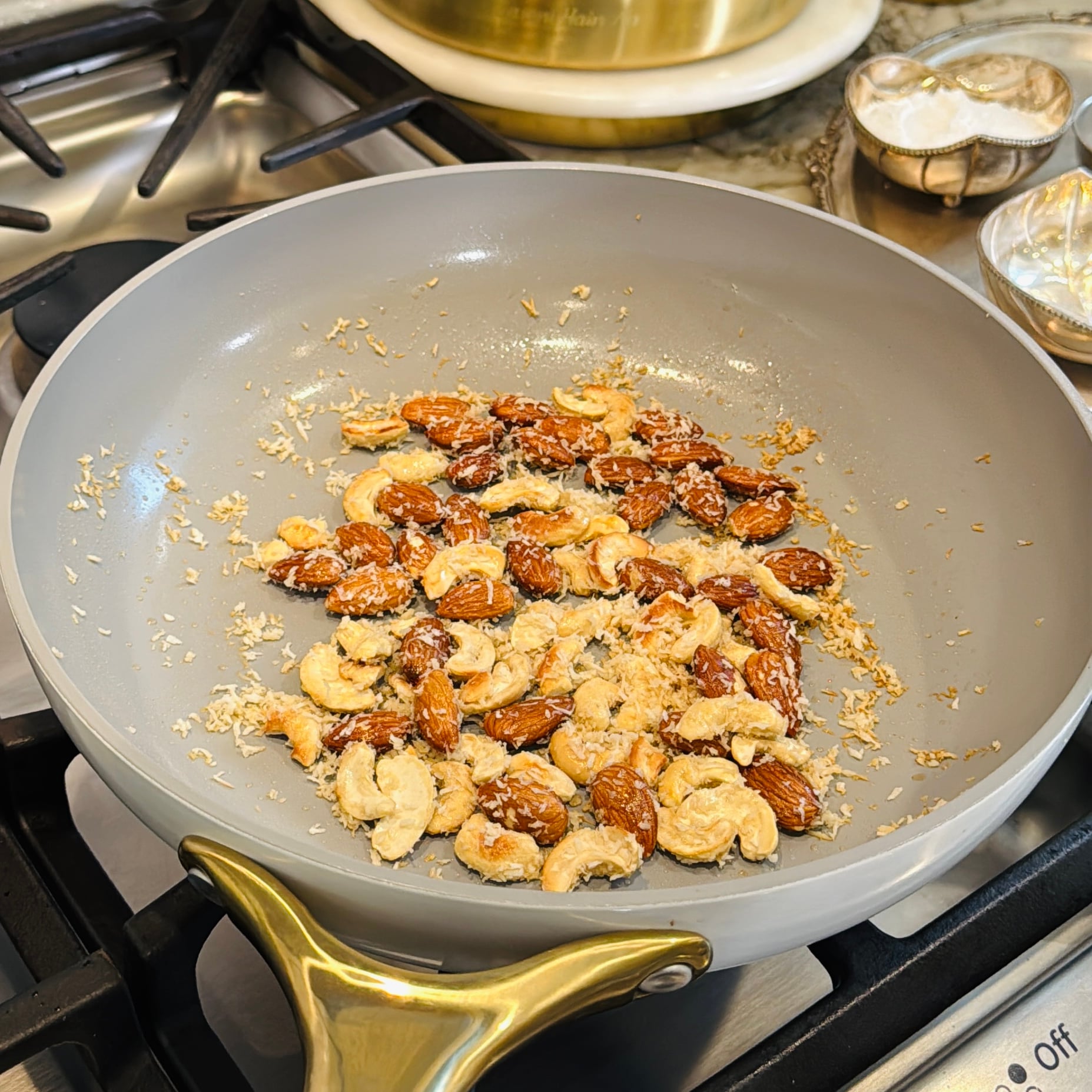 Almonds, cashews, and shredded coconut being roasted in a light gray skillet on a stove.