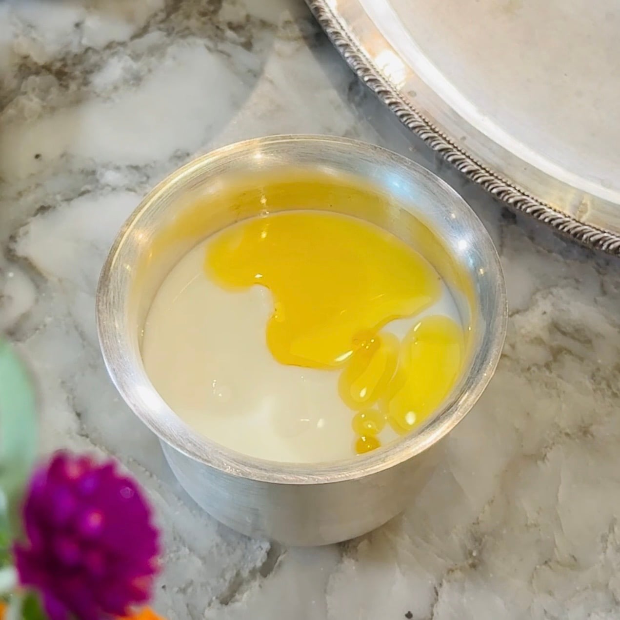 A metal cup containing milk and a cracked raw egg sits on a marble surface near a silver tray and a blurred purple flower in the foreground.