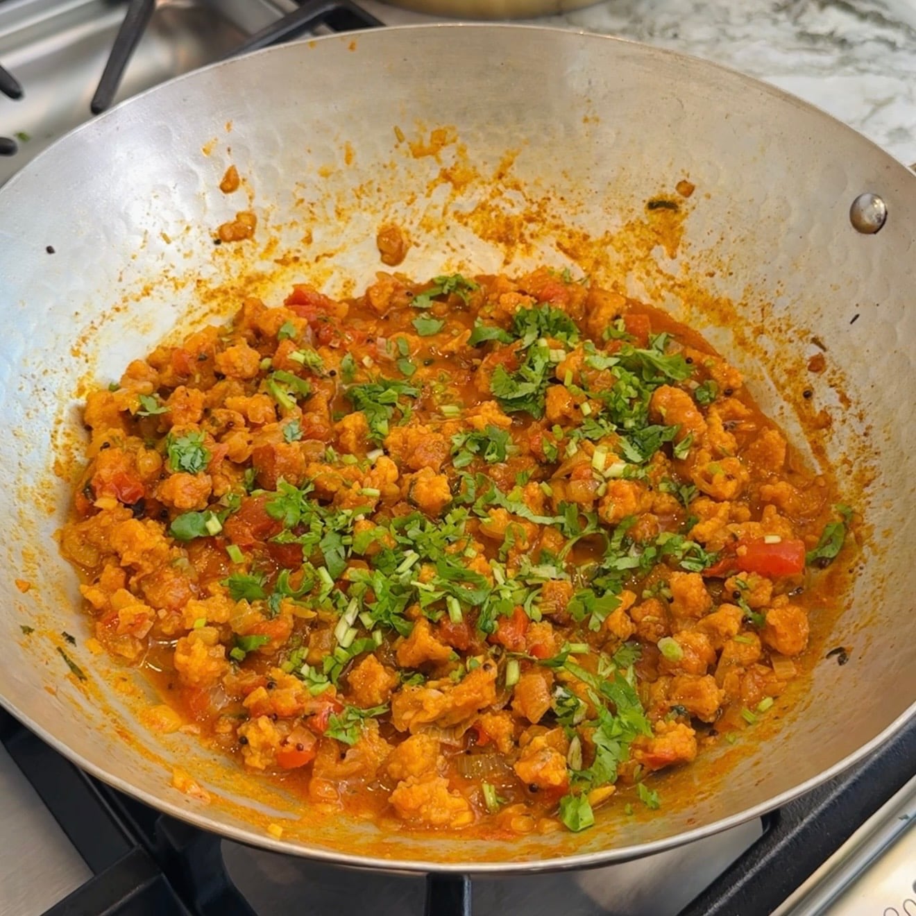 A stainless steel pan on a stove contains a cooked, sandge vegetable curry garnished with chopped cilantro.