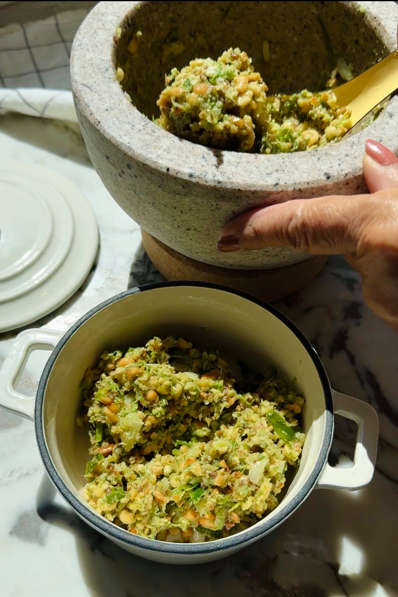 A hand holds a stone mortar filled with a chunky vegetable mixture, with some already served in a small white pot on a marble surface.