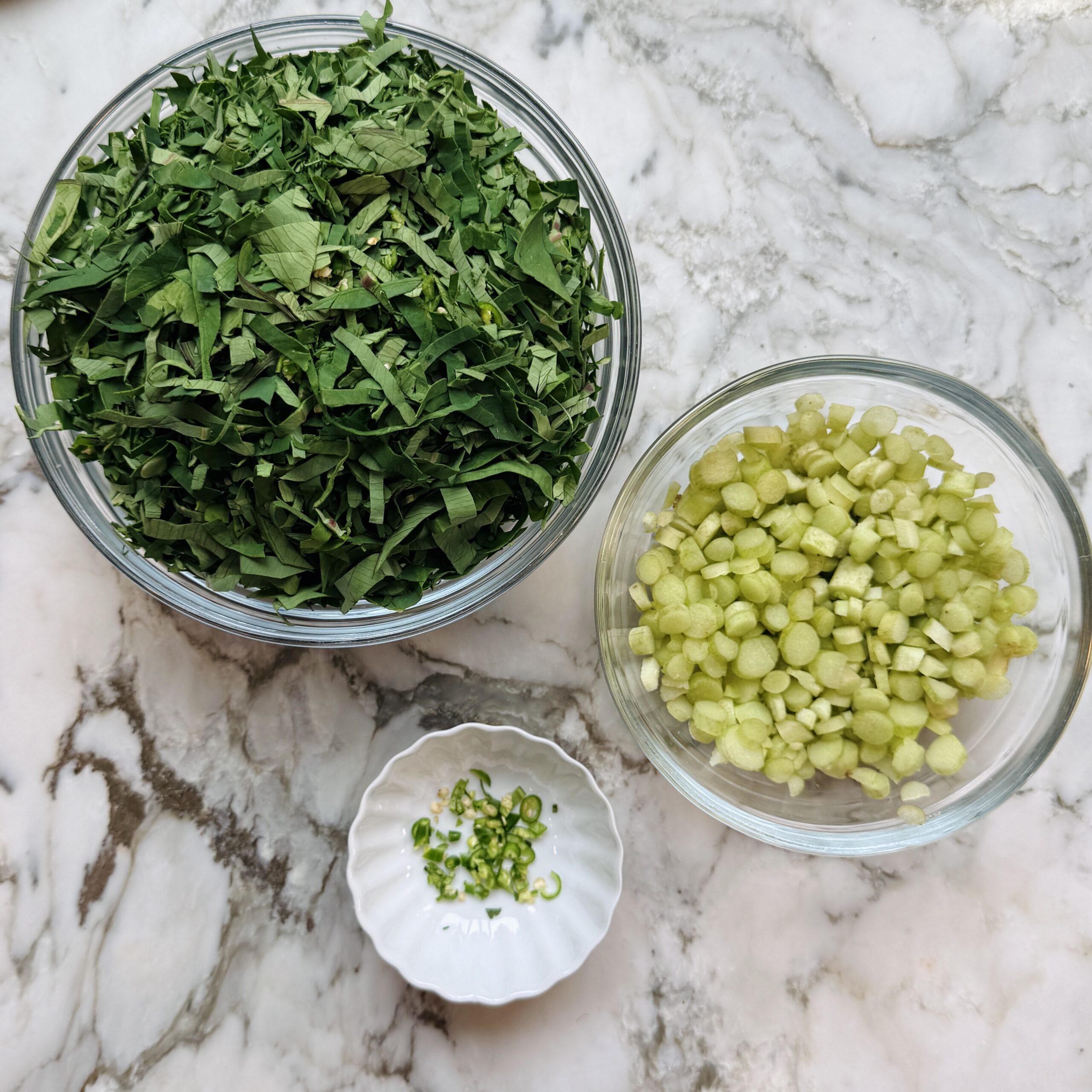 A glass bowl of chopped colocasia leavess, a glass bowl of diced stems, and a small dish of sliced green chili peppers.