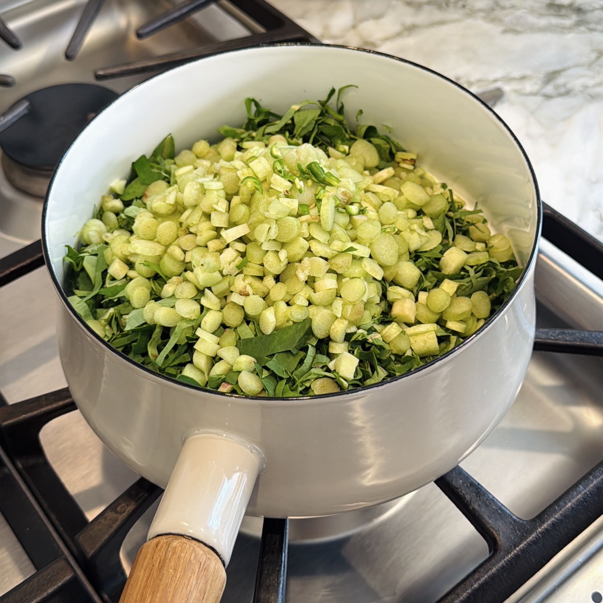 A light-colored pot on a stovetop filled with chopped leafy greens and sliced Colocasia stalks.