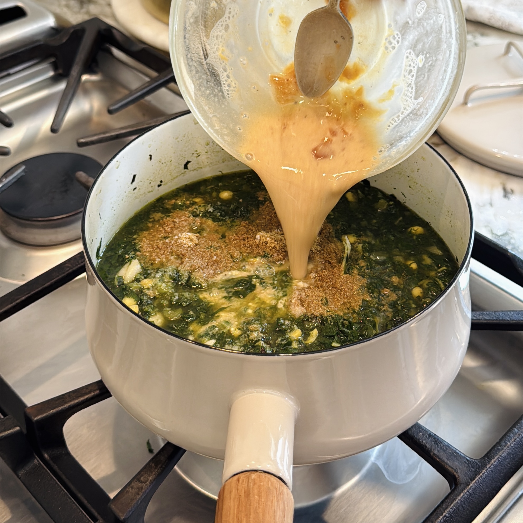 A hand pours a besan, tamarind and jaggery mixture from a glass bowl into a pot of simmering green leafy curry on a stovetop.