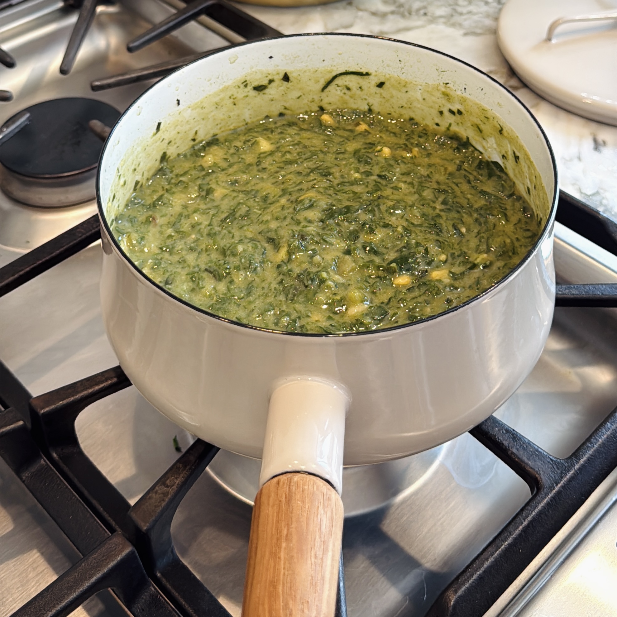 A light-colored saucepan on a gas stovetop filled with creamy green alu chi bhaji.