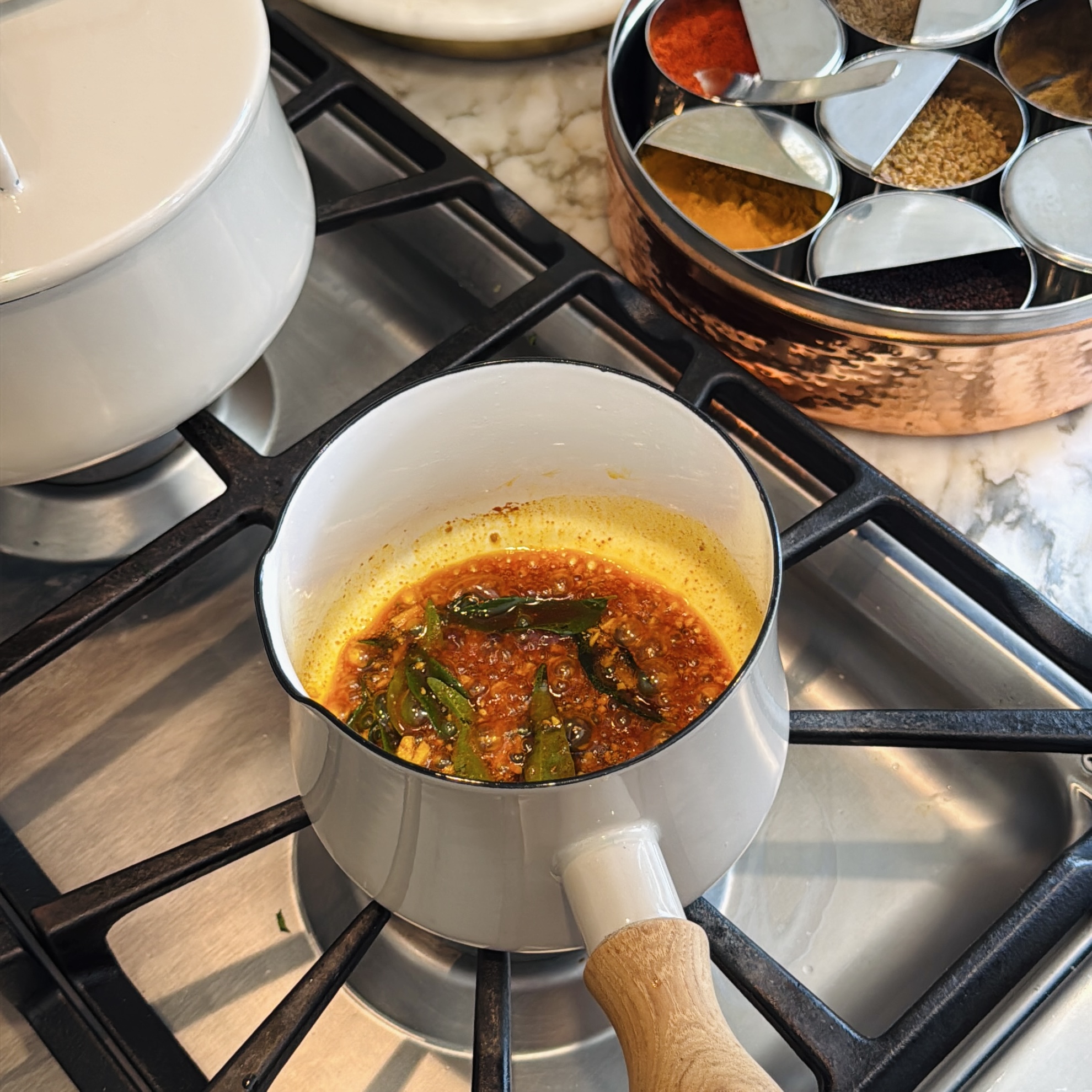 Spices and curry leaves frying in oil in a saucepan on a stovetop, with a spice tin nearby.