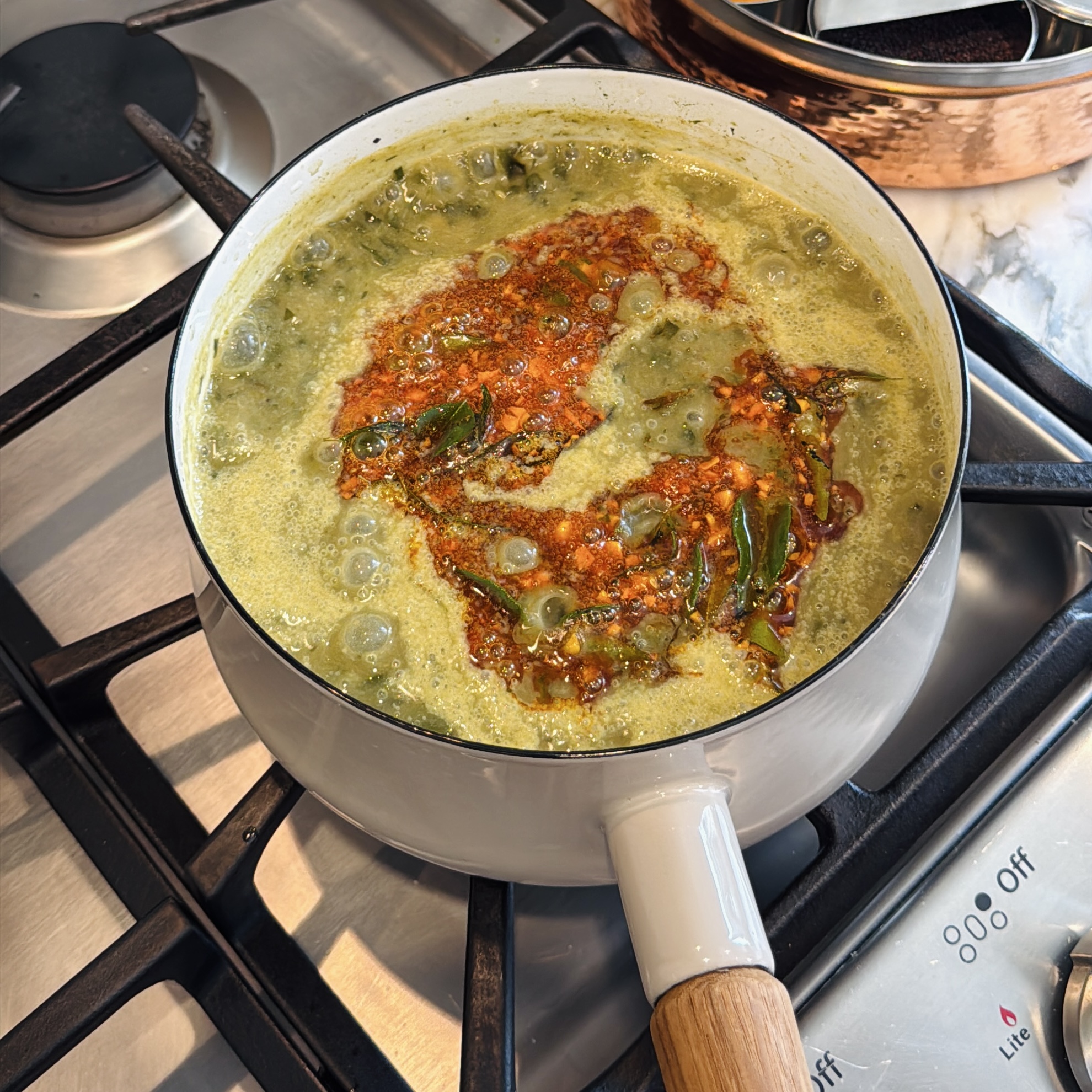A light-colored saucepan on a gas stovetop containing a simmering green curry topped with a tadka drizzle.