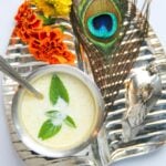 A metal tray with a bowl of yogurt garnished with mint, a spoon, marigold flowers, a peacock feather, and a small silver figurine.