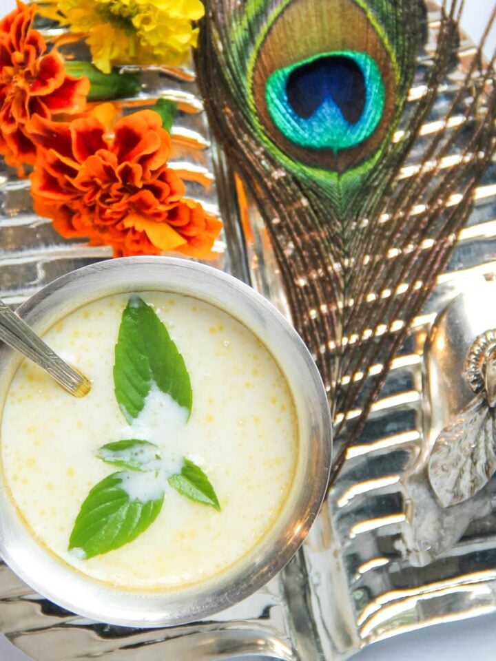 A metal tray with a bowl of yogurt garnished with mint, a spoon, marigold flowers, a peacock feather, and a small silver figurine.