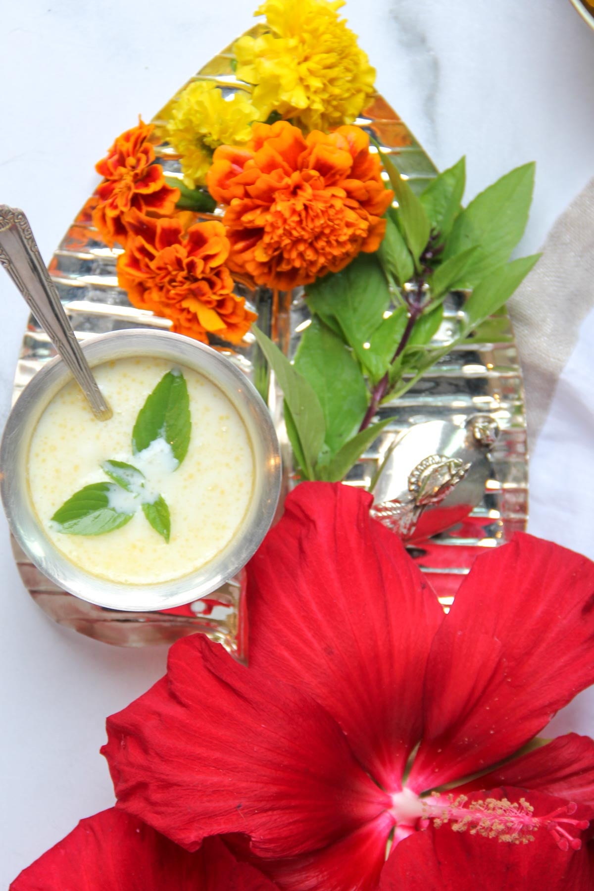 A silver tray with marigold and hibiscus flowers, green leaves, and a metal cup of creamy panchamrit garnished with holy basil.