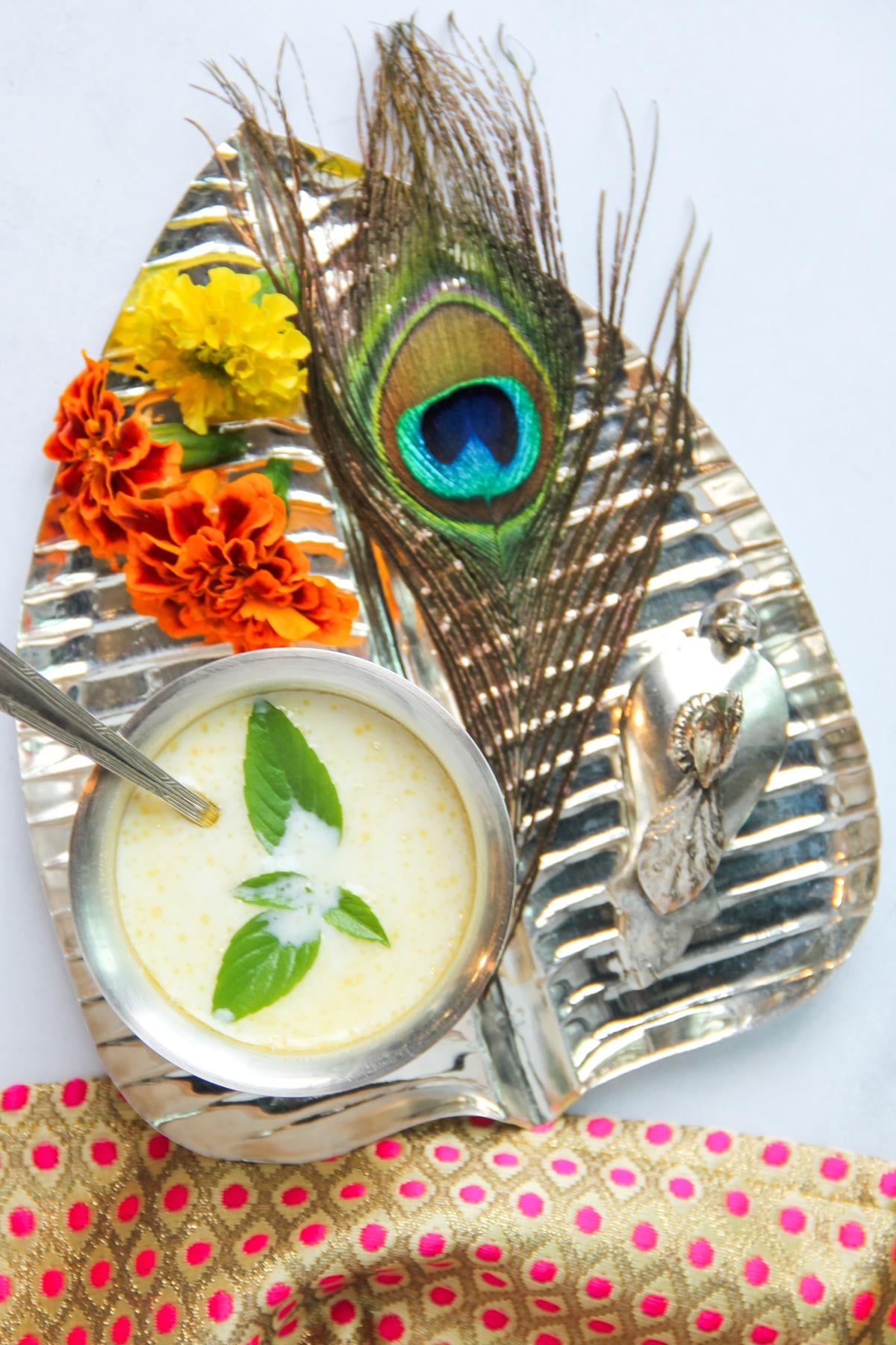 A silver tray with marigold flowers, a peacock feather, a metal Krishna idol, and a cup of panchamrut garnished with tulsi leaves, placed on a white background with a pink polka dot cloth.