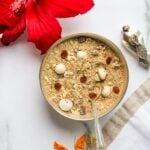 A bowl of panjiri beside a red hibiscus flower and a silver spoon on a marble surface.