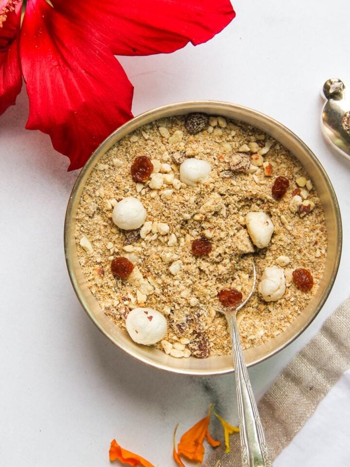 A bowl of panjiri beside a red hibiscus flower and a silver spoon on a marble surface.