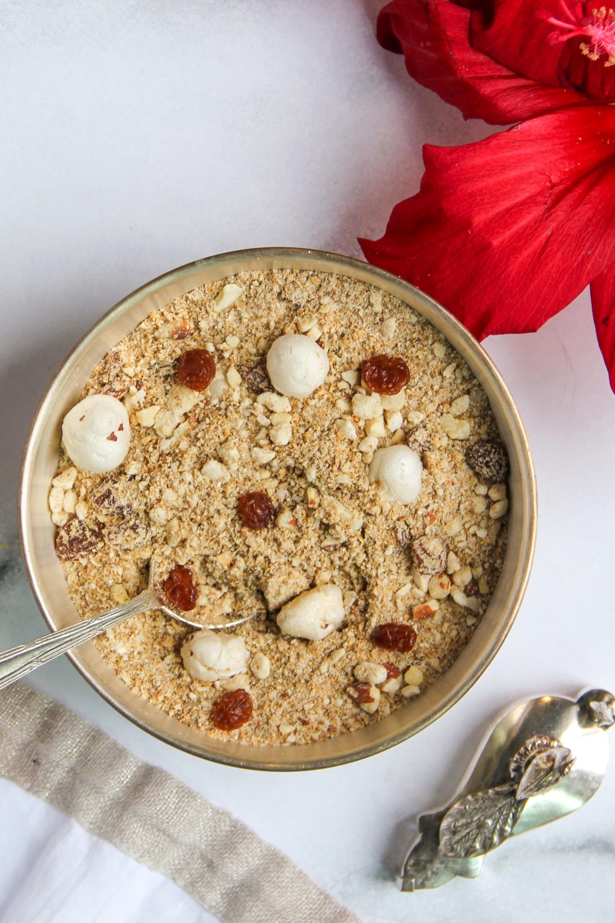 A bowl of panjiri with makhana and raisins, a spoon inside, red flower petals nearby.