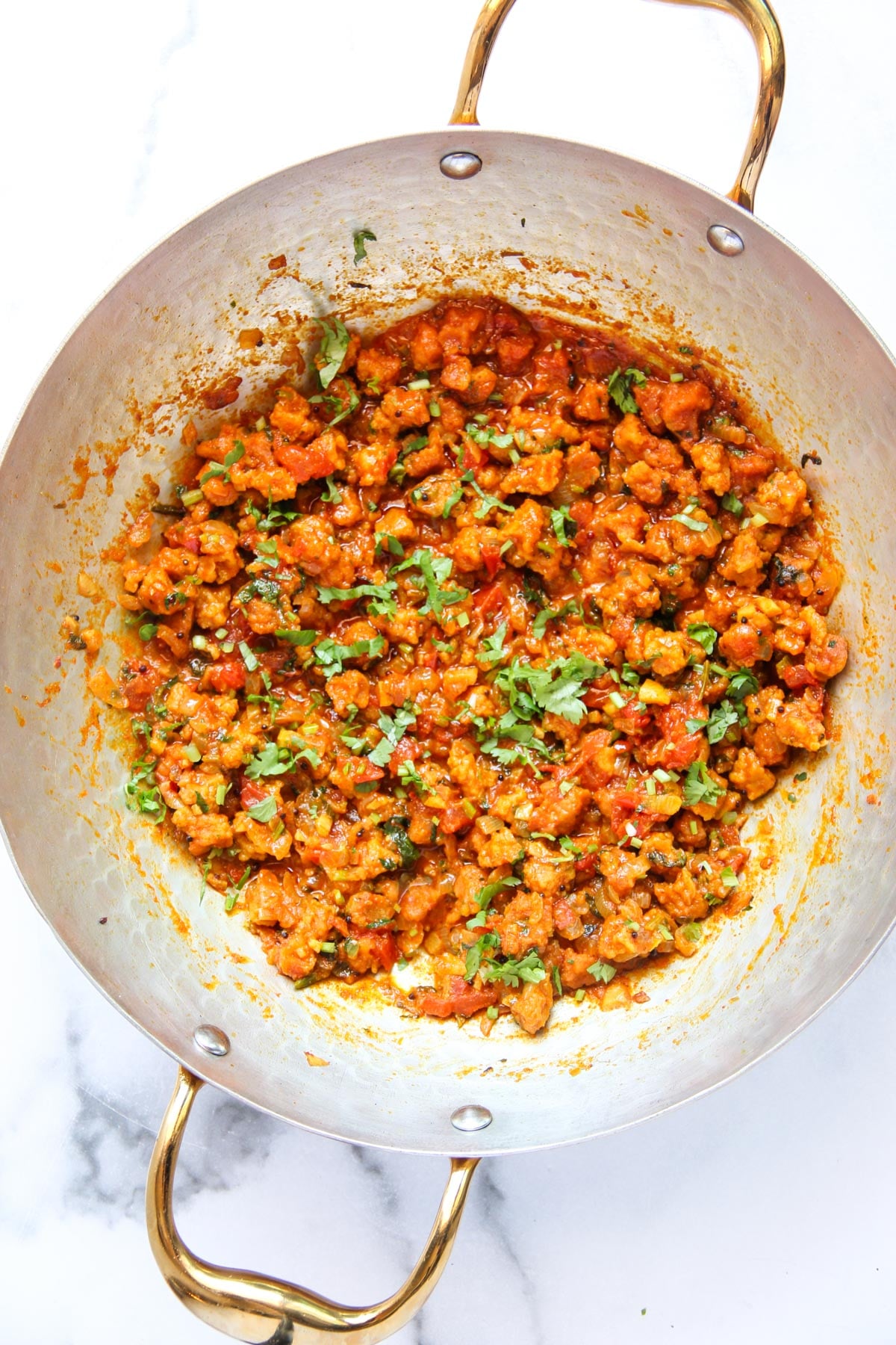 A metal kadai with sandge curry in a tomato-based sauce, garnished with chopped cilantro, on a white surface.