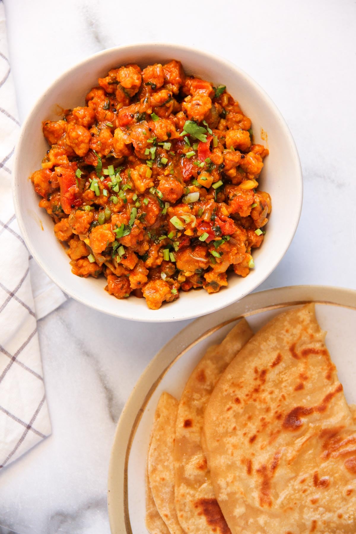A bowl of cooked vegetable curry garnished with chopped herbs, next to a plate with several pieces of flatbread on a marble surface.