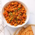 A bowl of cooked sandge bhaji garnished with cilantro, next to a plate with chapati on a white surface.