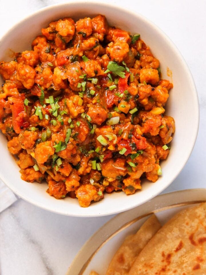 A bowl of cooked sandge bhaji garnished with cilantro, next to a plate with chapati on a white surface.