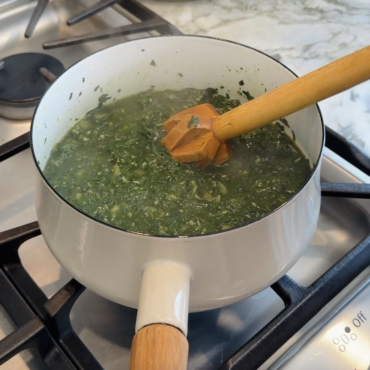 A white pot on a stovetop filled with chopped green vegetables being stirred with a wooden whisk.