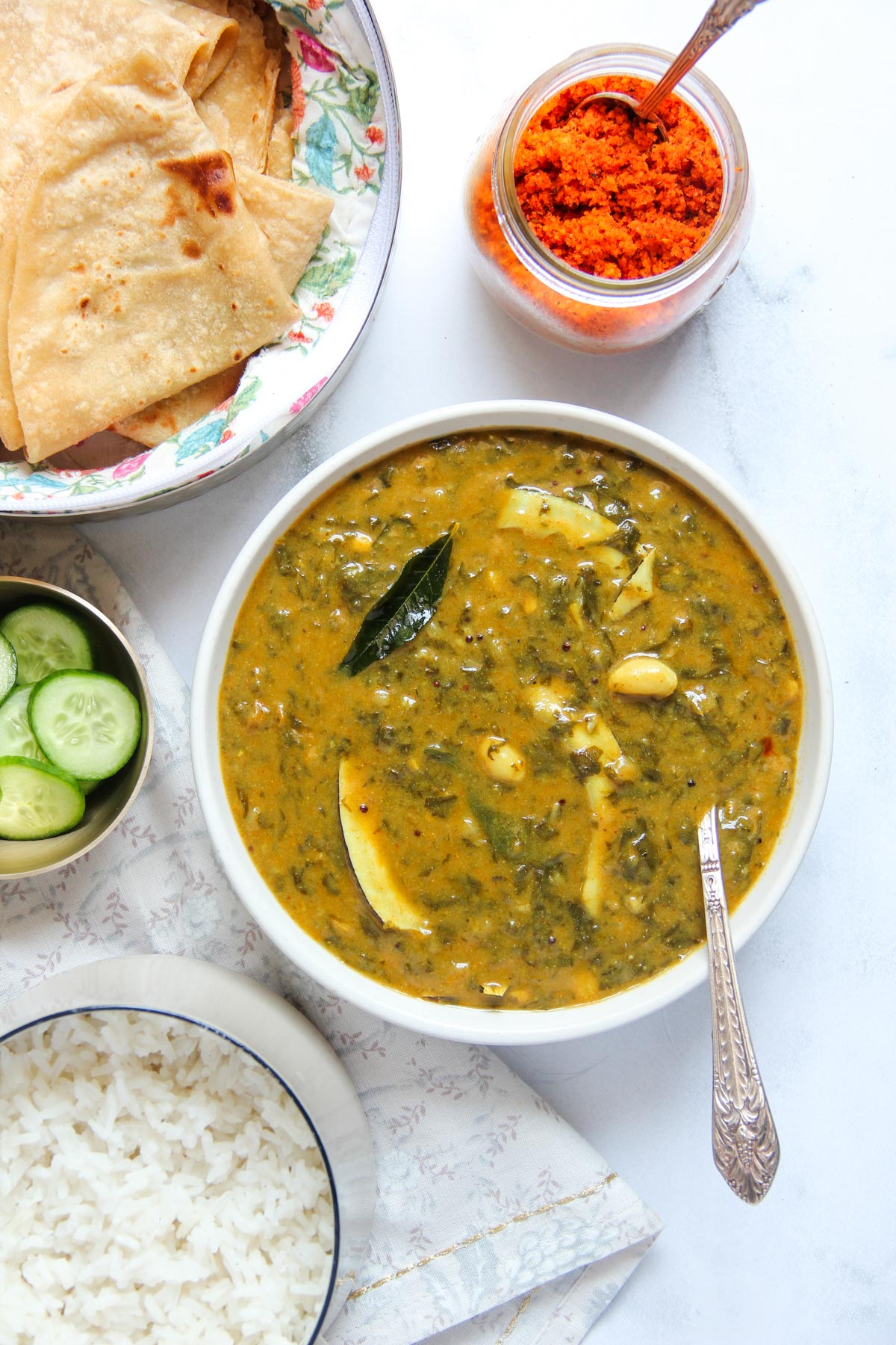 A bowl of alu chi bhaji, served with chapati, rice, sliced cucumbers, and red chutney.
