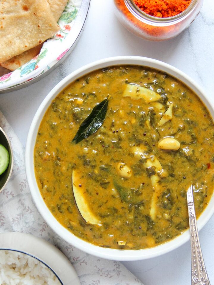 A bowl of leafy green vegetable curry with sliced potatoes, served with roti, rice, and cucumber slices.