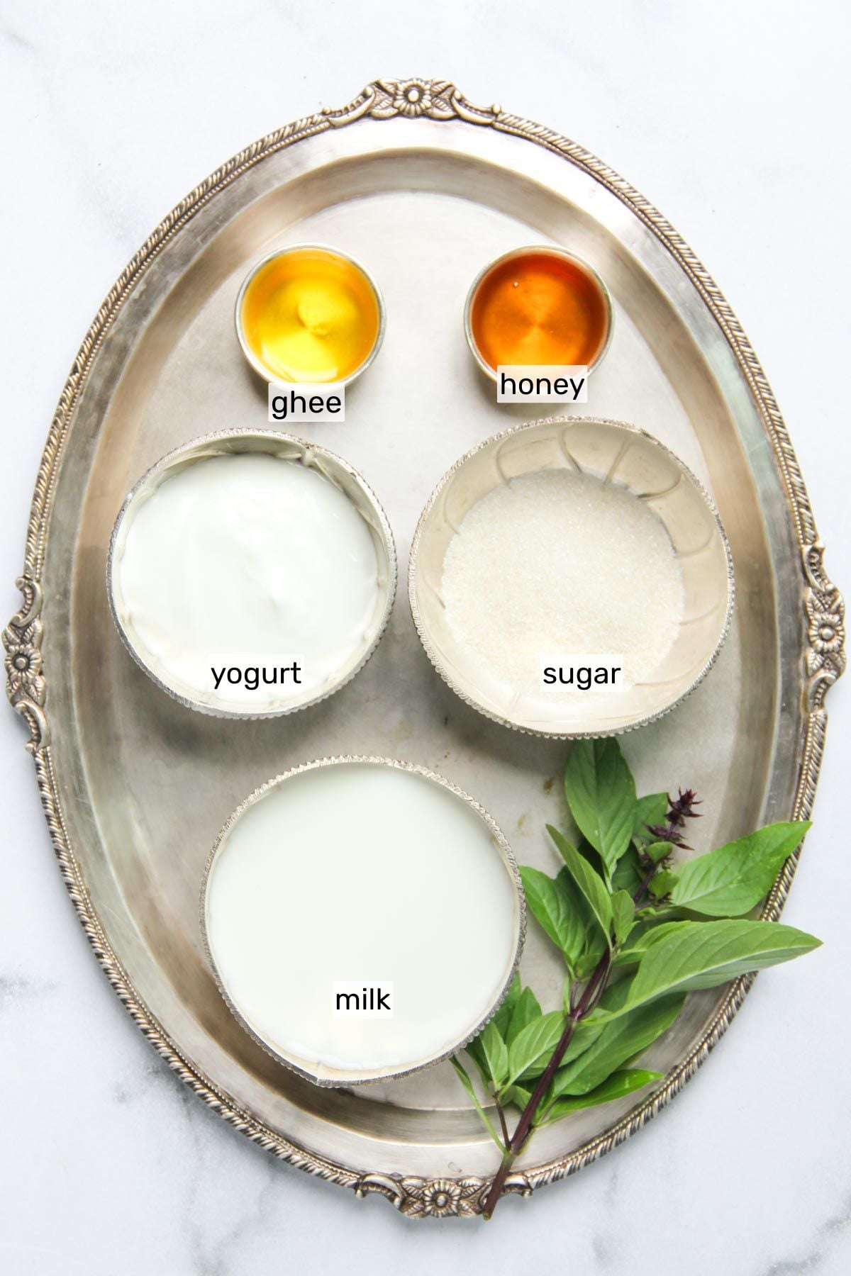 A silver tray holds labeled bowls of ghee, honey, yogurt, sugar, and milk, along with sprigs of fresh holy basil on a white background.
