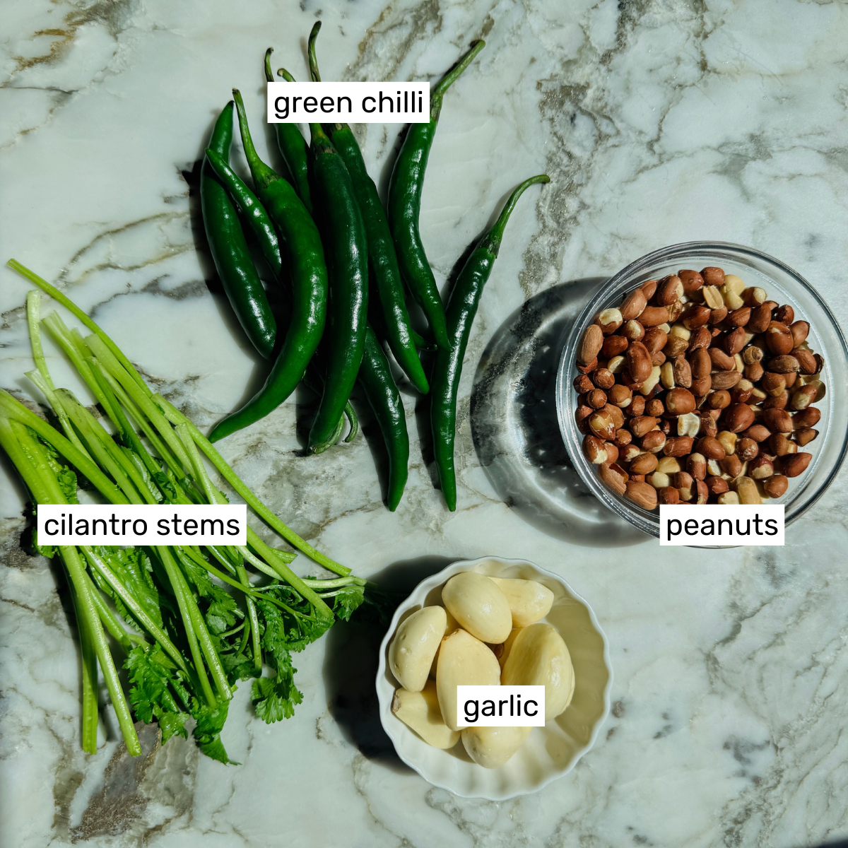 Green chillies, cilantro stems, peanuts in a glass bowl, and peeled garlic cloves in a small dish on a marble surface, each labeled with text.