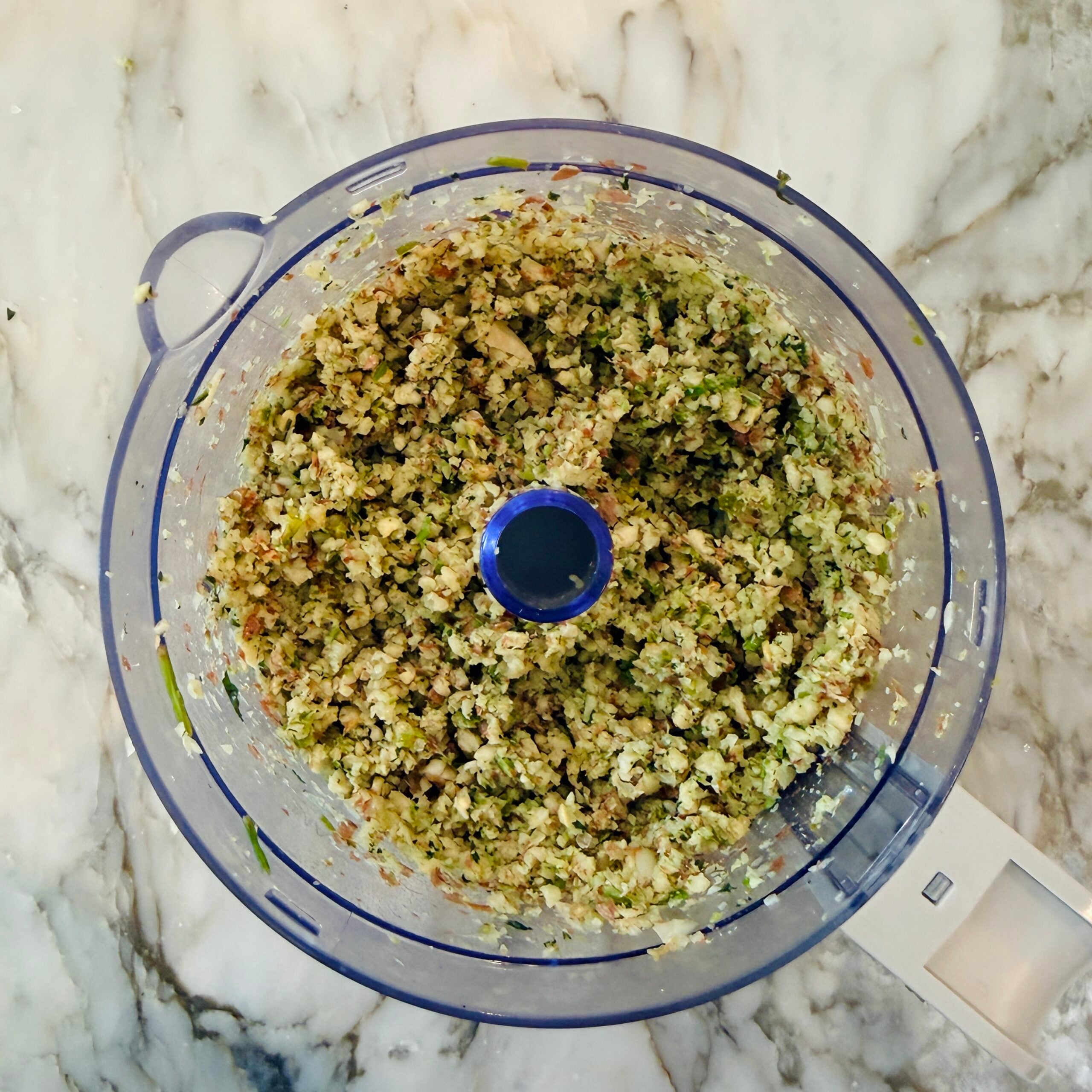 Food processor bowl filled with finely chopped vegetables and nuts on a marble countertop.