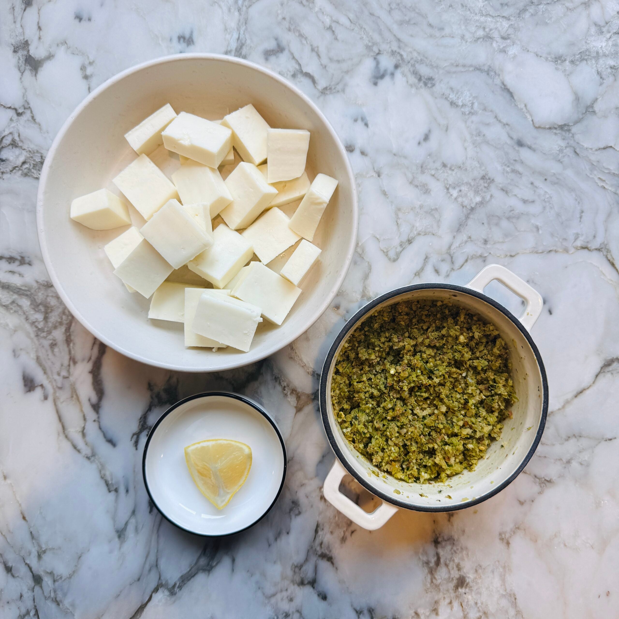 A bowl of cubed paneer, a bowl of herb paste, and a lemon wedge on a marble surface.