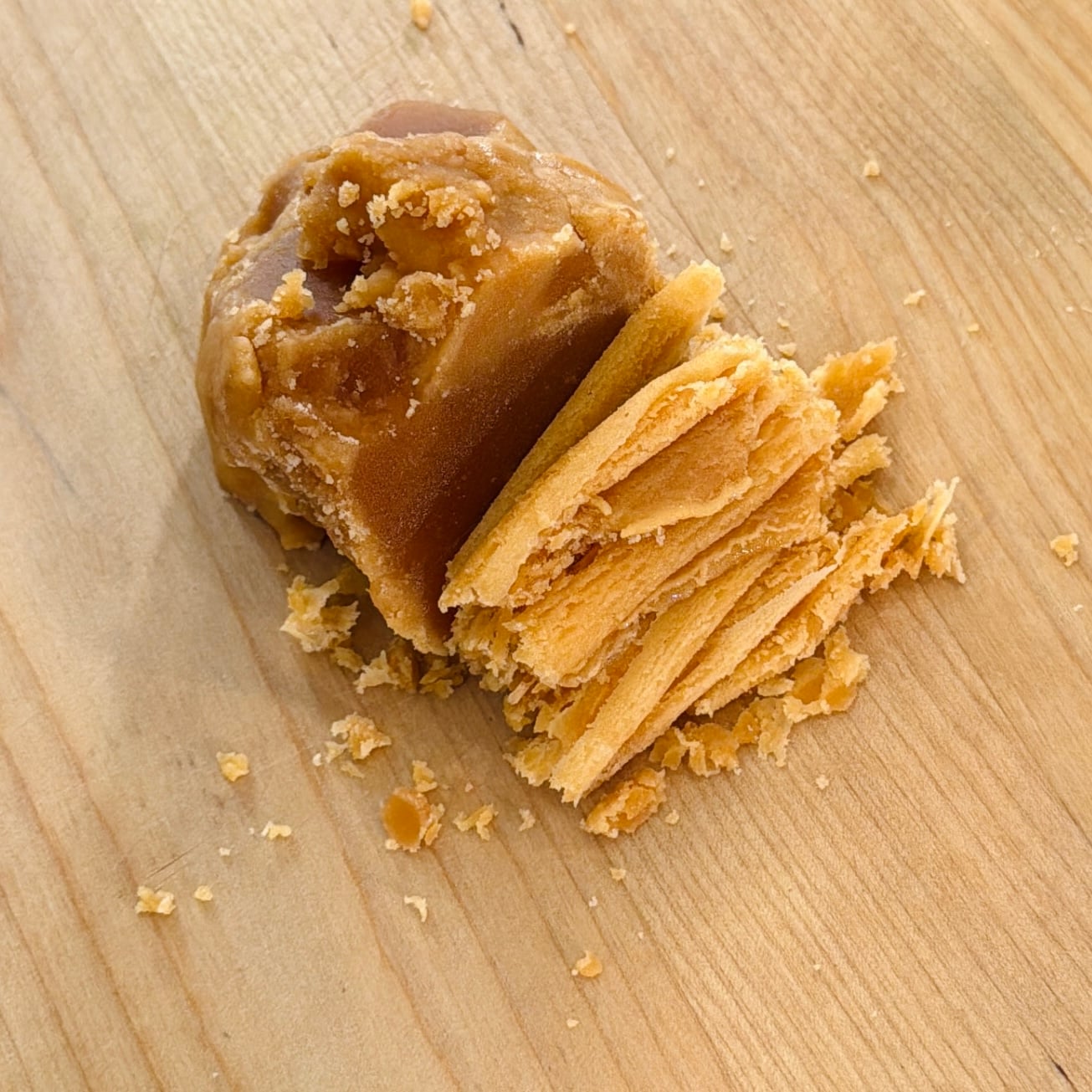 A block of brown jaggery with a portion shaved into thin, layered pieces on a wooden surface.