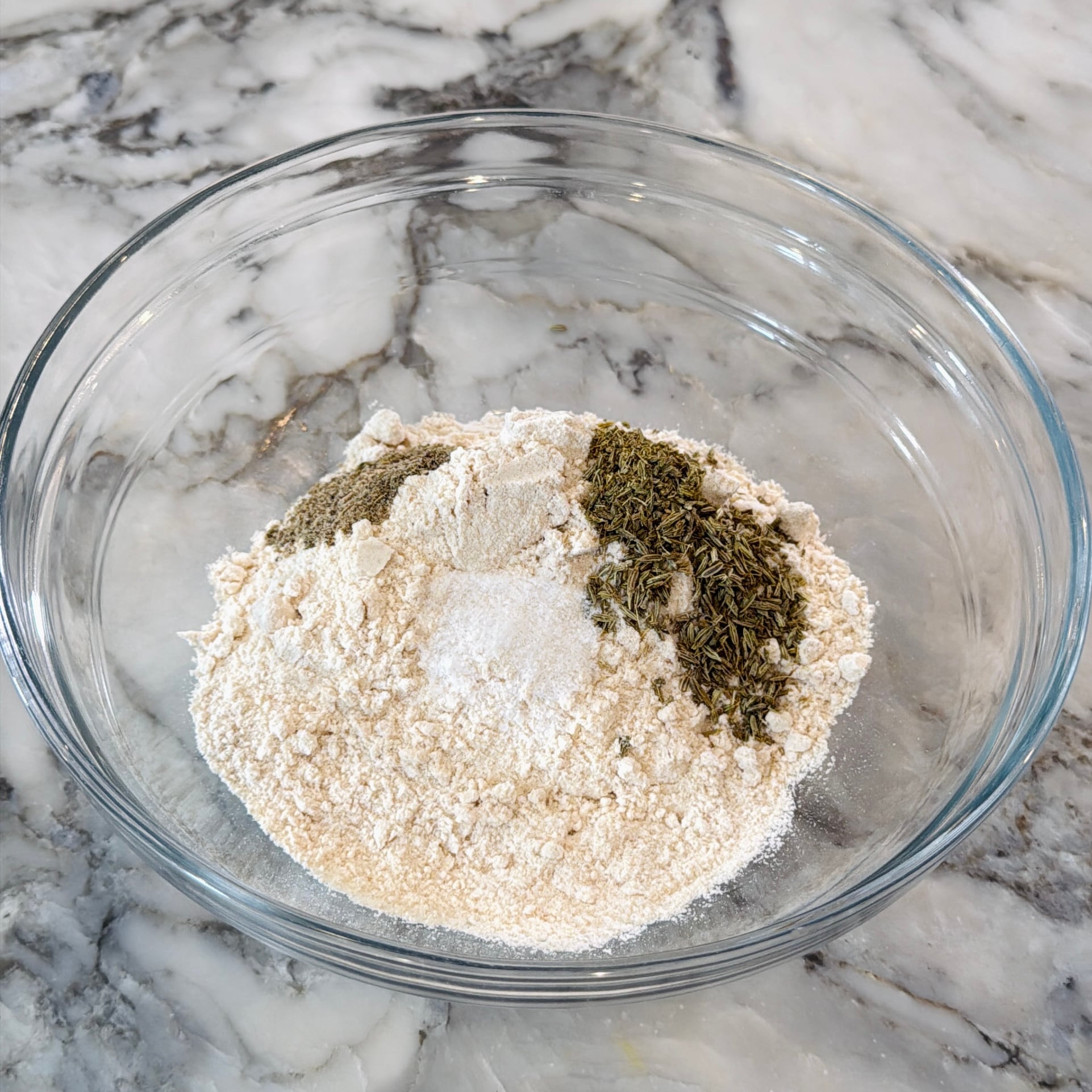 A glass bowl containing a pile of flour topped with salt and dried herbs.