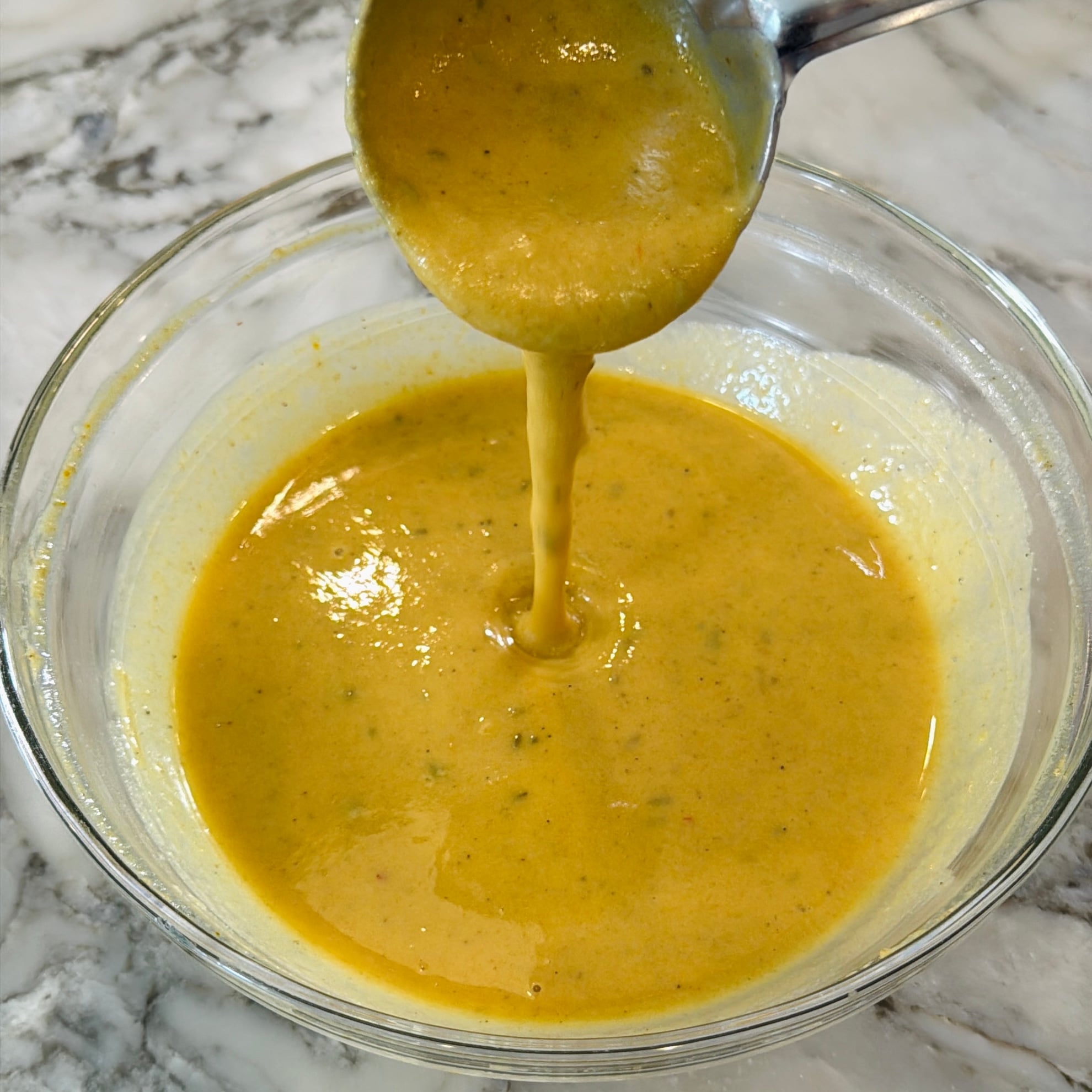 A metal ladle pouring yellow sauce into a glass bowl.