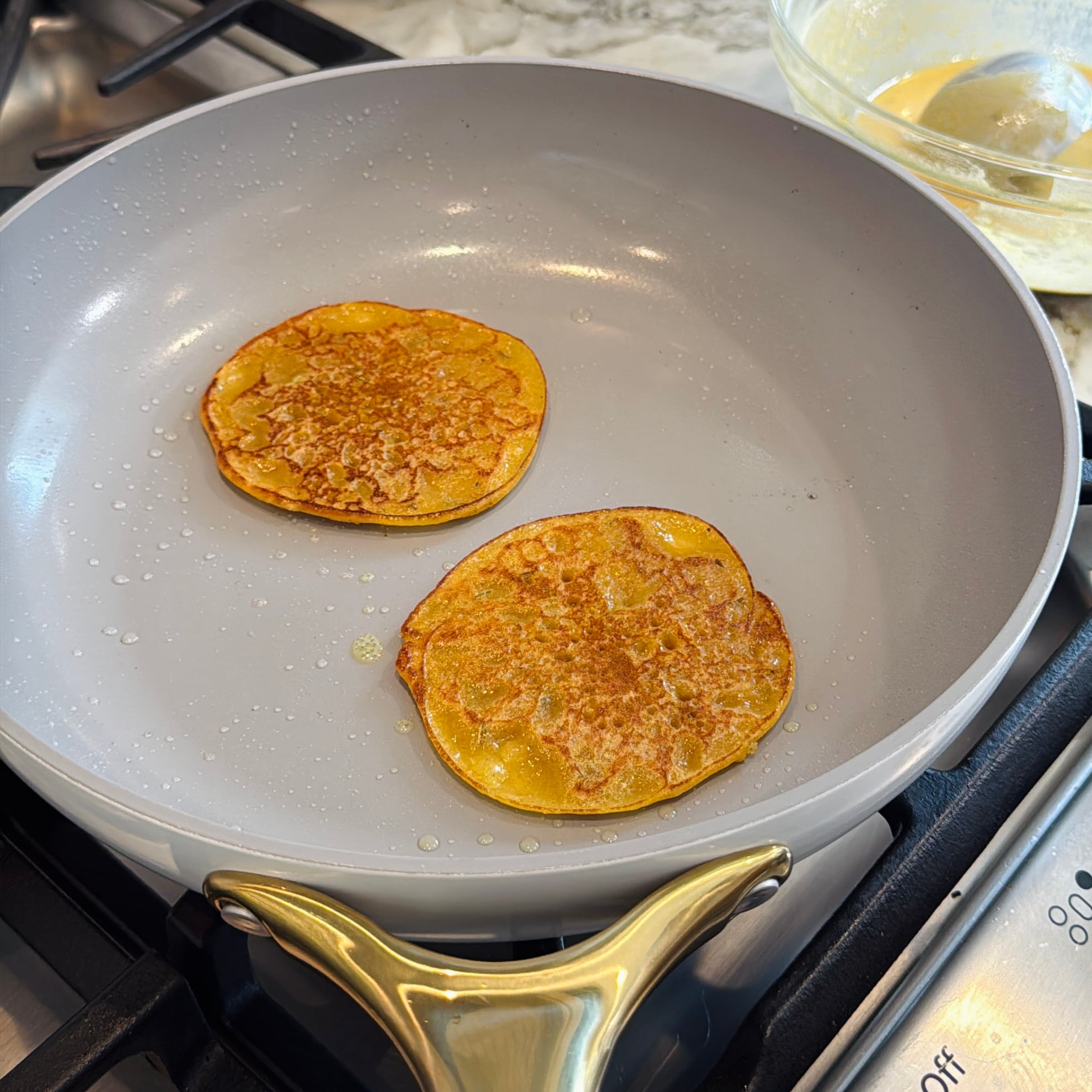 Two pancakes cooking in a grey nonstick skillet on a stovetop.