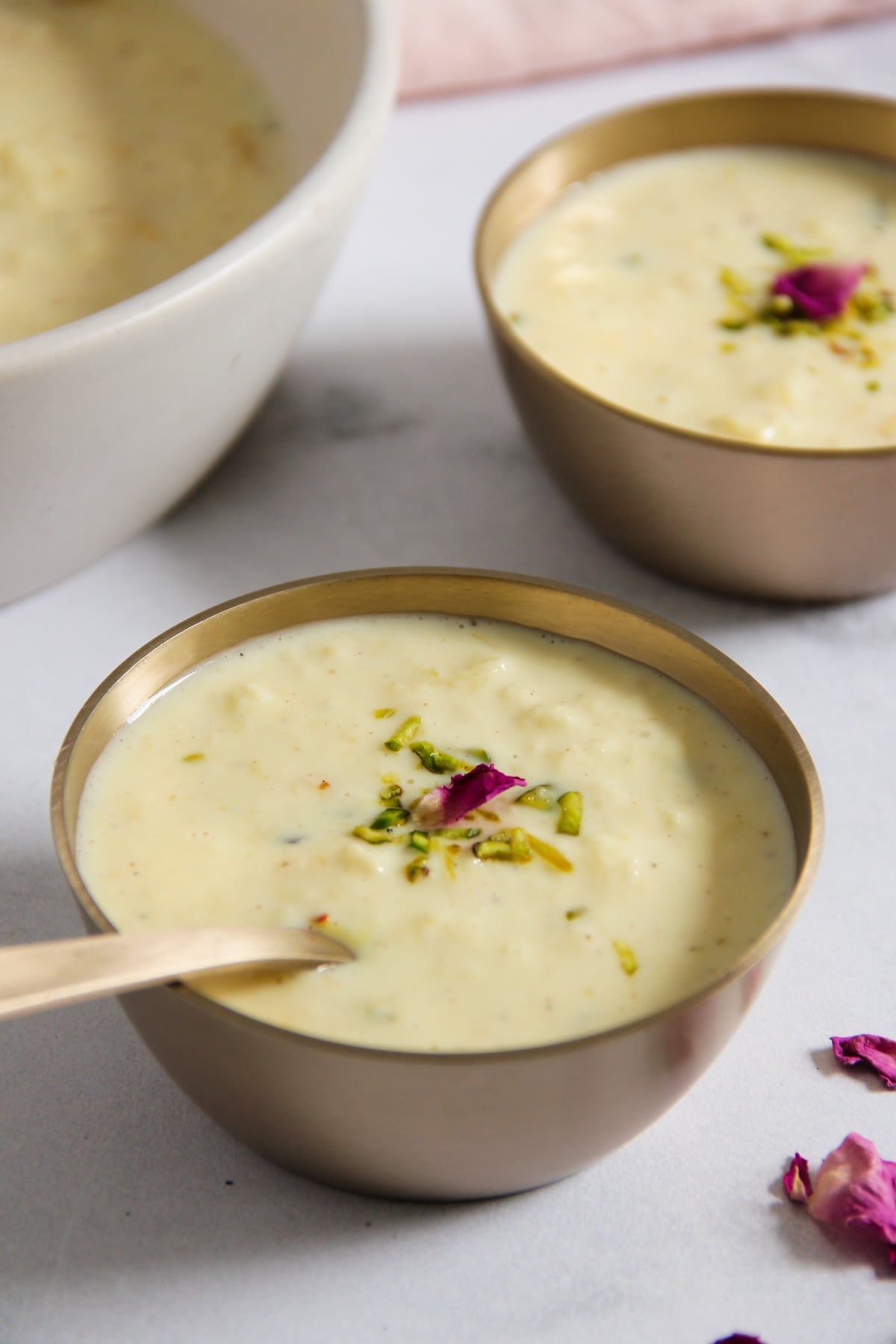 Two brass bowls filled with creamy randi, garnished with chopped pistachios and rose petals.